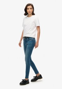 T-shirt blanc à col rond, jeans skinny bleus et mocassins noirs avec détail en nœud. Le mannequin se tient sur un fond neutre, mettant en valeur la tenue.