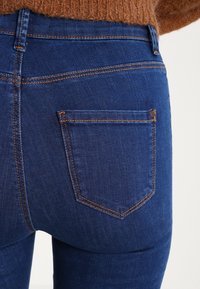 Dark blue denim jeans featuring a back pocket with orange stitching detail; smooth texture with a slight sheen; fitted design.