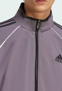 Siva poliester jakna na zip z visokim ovratnikom, ki ima črne detajle in tri črne črte po ramenih; logotip Adidas na prsih.