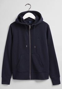 Mörkblå zip hoodie med framfickor och dragsko på huvan, hängande på en vit galge mot en grå bakgrund.