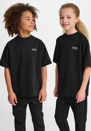 Deux enfants se tiennent côte à côte, portant des chemises et des pantalons noirs à manches courtes assortis, se souriant l'un à l'autre sur un fond blanc.