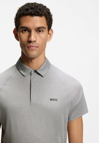 Mand iført en grå kortærmet polo med "BOSS"-logo på venstre bryst, set forfra mod en hvid baggrund.