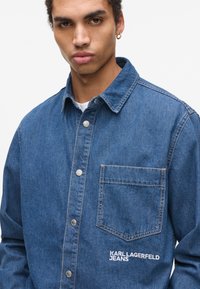 Traper košulja u srednjem plavom s dugim rukavima, dugmadi sprijeda, šiljastim ovratnikom, džepom na prsima i bijelim vezom s brendom na džepu.