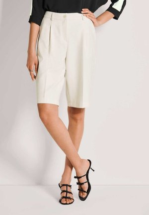 Femme portant un short blanc sur mesure jusqu'aux genoux, un haut noir et des sandales noires à talons avec brides, debout, jambes croisées.