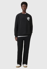 Schwarzes Sweatshirt mit einem weißen Grafikdruck auf der Brust, kombiniert mit schwarzen Hosen und weißen Sneakers. Mit Rundhalsausschnitt und langen Ärmeln.