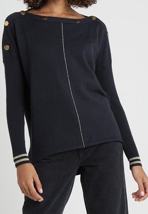 Kvinde iført en mørkeblå sweater med guldknapper på skuldrene, lodret syning foran og stribede manchetter, kombineret med sorte jeans.