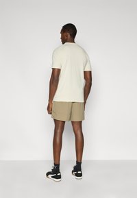 Ανοιχτόχρωμο κρεμ κοντομάνικο μπλουζάκι, χακί σορτς, μαύρα αθλητικά παπούτσια, όρθιος από το πίσω μέρος με απλό φόντο.