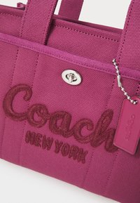 Purple canvas tote bag featuring embroidered "Coach New York" text, silver hardware, and a small branded tag attached by a chain.