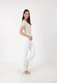 Top ajustado beige con mangas cortas, combinado con pantalones blancos de campana de cintura alta. La modelo está de perfil, mostrando unos zapatos de tacón minimalistas con tiras.