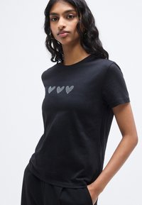 Μαύρο κοντομάνικο μπλουζάκι από απαλό ύφασμα με τρεις καρδιές διακοσμημένες με στρας στο στήθος. Ραμμένη εφαρμογή.