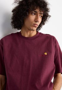 Burgundy cotton t-shirt with a crew neckline, short sleeves, and an embroidered logo in yellow on the left chest area. Smooth texture.
