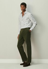 Valge särk, millel on värvikas lillemuster, kombineeritud oliivroheliste lõngveluurpükstega. Tumepruunid libisemisjalatsid, seisab neutraalse tausta ees.