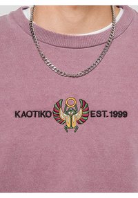 Un uomo che indossa una collana d'argento e un felpa viola con un scarabeo colorato ricamato e il testo "KAOTIKO EST. 1999".
