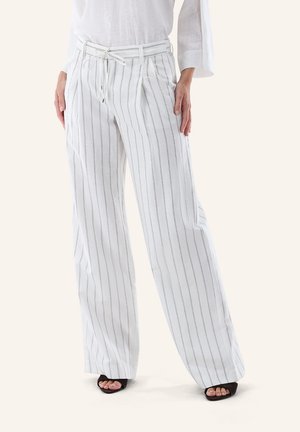 Frau trägt weiße, weit geschnittene Hose mit dünnen vertikalen grauen Streifen und einem Kordelzug am Bund, kombiniert mit schwarzen, offenen Sandalen mit Absatz.