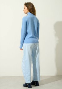 Helles blaues Wollpullover mit hohem Kragen, kombiniert mit hellblauenweiten Hosen; das Model trägt marineblaue Plateauschuhe. Glatte Textur, lässige Passform.