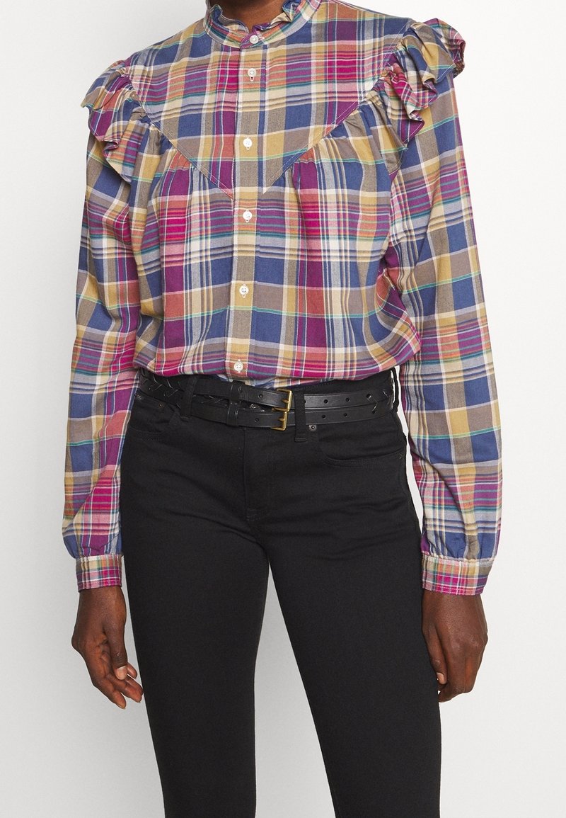 Femme portant une chemise à carreaux multicolore boutonnée avec des épaules à volants, rentrée dans un pantalon noir taille haute avec une ceinture noire.