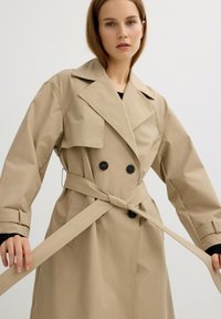 Abrigo trench beige con grandes solapas, frente de doble botonadura, botones negros y cinturón atado. Presenta mangas largas y una textura suave y brillante.
