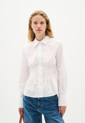 Weißes Baumwollhemd mit klassischem Kragen, vorderen Knöpfen und vertikalen Falten an der Taille. Lange Ärmel und eine figurbetonte Silhouette, kombiniert mit blauen Jeans.