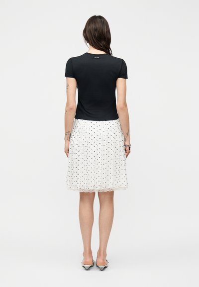 Γυναίκα με μακριά σκουρόχρωμα μαλλιά φοράει μαύρο μπλουζάκι με κοντά μανίκια και λευκή φούστα μέχρι τα γόνατα με μικρές μαύρες βούλες, στέκεται με την πλάτη στραμμένη.