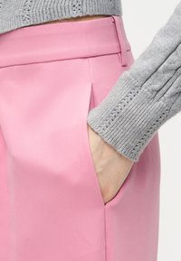 Χέρι στην τσέπη μιας ροζ φούστας με ψηλή μέση, συνδυασμένη με μανίκι γκρι πλεκτού πουλόβερ με σχέδιο καλωδίου.