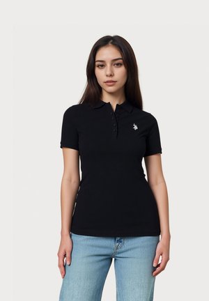 Tânără purtând un tricou polo negru mulat cu mânecă scurtă și logo pe piept și blugi albastru deschis, stând în picioare pe un fundal simplu.