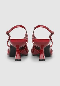 Rote Sandaletten aus Lackleder mit hohem Absatz, abgerundeter Spitze, Knöchelriemen und dickem, konischem Absatz. Glatte Textur und glänzende Oberfläche.