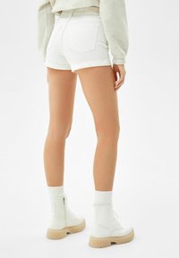 Mujer vestida con pantalones cortos blancos con dobladillos enrollados, suéter corto beige, calcetines blancos y botas blancas con cordones y suelas beige, de pie sobre un fondo blanco.