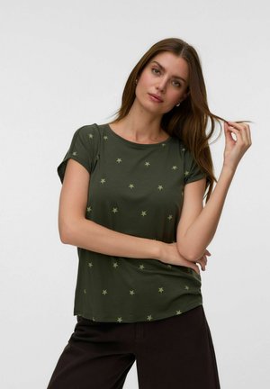 Femme aux longs cheveux bruns porte un t-shirt vert olive avec de petits motifs d'étoiles, tenant une mèche de cheveux et regardant la caméra.
