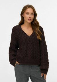 Pull en maille épaisse marron foncé avec un col en V, présentant des motifs de torsades texturés, des poignets et un ourlet côtelés. Le tissu semble doux et confortable.