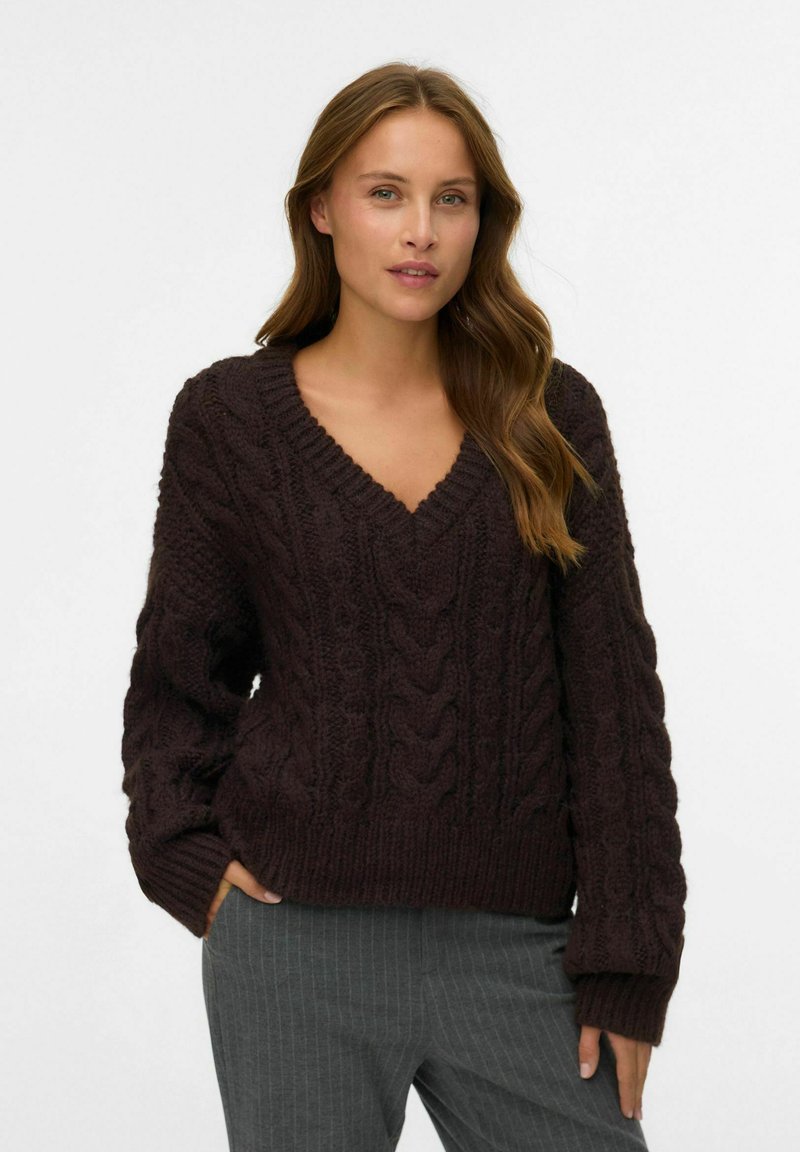 Pull en maille épaisse marron foncé avec un col en V, présentant des motifs de torsades texturés, des poignets et un ourlet côtelés. Le tissu semble doux et confortable.
