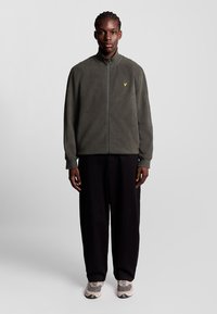 Fleecejacka i mörkgrön med hög krage, hel dragkedja och liten gul logotypdetalj; ihop med löst svart byxa och sneakers.