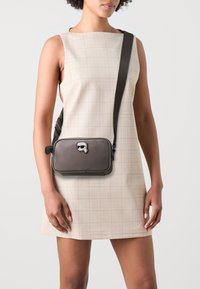 Borsa a tracolla rettangolare grigia scura realizzata in materiale liscio, con un accento del logo e una tracolla robusta. Indossata sopra un vestito a quadri chiari.