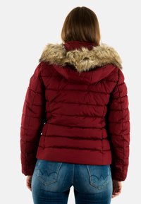 Bordeauxrode donsjas met een capuchon van nepbont, voorzien van doorgestikte delen en een aangesloten ontwerp. Gedragen over een blauwe spijkerbroek met een decoratieve achterzak.