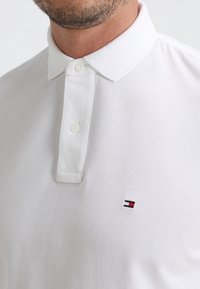 Polo shirt blanc avec un tissu texturé, un col classique et trois boutons. Présente un petit logo rouge, blanc et bleu sur la poitrine.