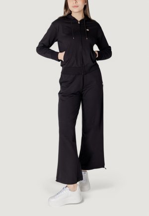 Haut à capuche zippé noir avec une poche avant, associé à un pantalon noir à jambe large. Les deux sont en tissu doux ; présentent des accents en métal doré. Baskets blanches.