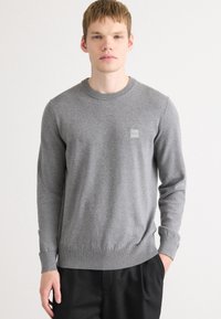Grauer Strickpullover mit langen Ärmeln, rundem Halsausschnitt, gerippten Bündchen und Saum, mit einem kleinen Markenpatch auf der Brust.