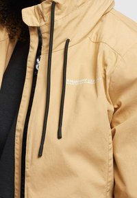 Chaqueta beige con capucha y cordones negros, usada sobre una camiseta negra con un pequeño texto blanco en el lado izquierdo del pecho.