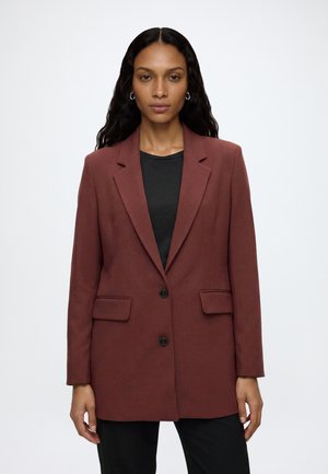 Femme aux longs cheveux ondulés portant un blazer rouge foncé boutonné par-dessus un haut noir sur un fond blanc uni.