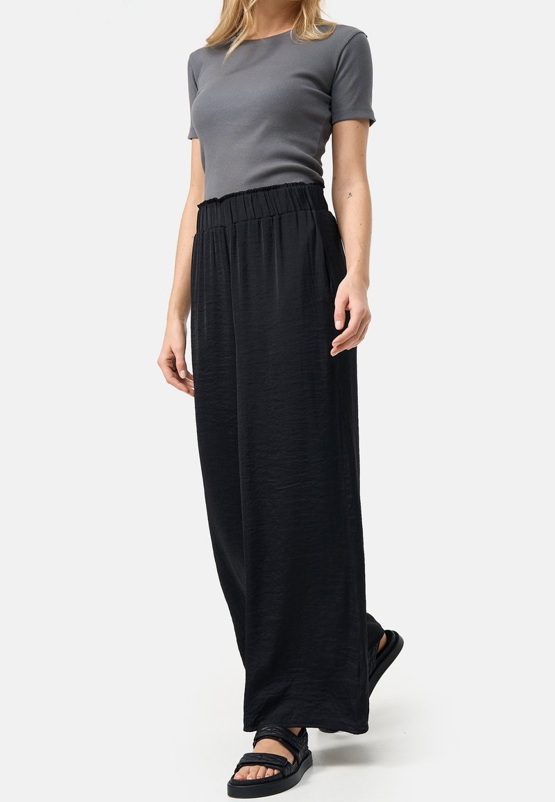 Femme portant un haut gris à manches courtes ajusté, un pantalon ample noir taille haute et des sandales noires, debout devant un fond blanc.