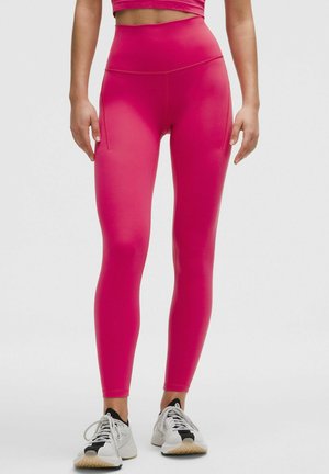 Skinnende lyserøde højtaljede leggings lavet af strækbart materiale, med sidelommer og en glat, elegant tekstur. Båret med hvide træningssko.