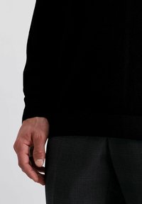 Schwarzer Pullover mit gerippten Bündchen, kombiniert mit dunkelgrauen Hosen, die ein feines Karomuster aufweisen. Der Fokus liegt auf der Hand, die auf der Taille ruht.