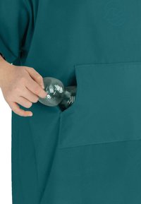 Ein türkisfarbenes Kleidungsstück mit einer großen Tasche wird gezeigt; eine Hand greift hinein, um eine transparente Plastikflasche mit gerippter Struktur zu fassen.