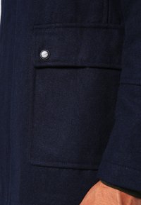 Marinblå ullkappa med en strukturerad finish, utrustad med en bröstficka med en silvertryckknapp och kontrasterande mörkgrönt trim på ärmen.