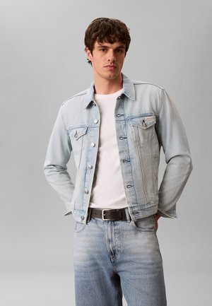 Młody mężczyzna ubrany w jasnoniebieską kurtkę jeansową na białą koszulę, z dopasowanymi jeansami i czarnym paskiem, stojący na jednolitym szarym tle.