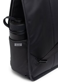 Bolsa mensajera negra hecha de material duradero con un bolsillo lateral que sostiene una botella de agua negra, con una etiqueta de marca y una correa ajustable.