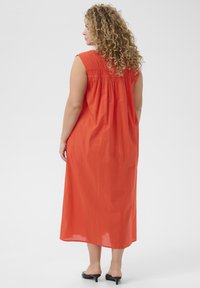 Langes, ärmelloses Kleid aus leuchtend orangefarbenem Stoff mit lockerer Passform und gerafftem Rückendetail. Besitzt einen geraden Saum und wird mit schwarzen Schuhen kombiniert.