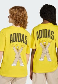 Camiseta de algodón amarilla con un personaje de dibujos animados, gran texto "ADIDAS" y un diseño juguetón de un personaje con atuendo beige, de mangas cortas.