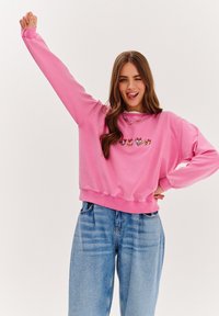 Różowa bluza z długim rękawem, z prążkowanymi mankietami i zaokrąglonym dołem, z kolorową grafiką kreskówkowych postaci na klatce piersiowej.
