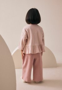 Top peplum cu carouri roz deschis, cu mâneci lungi și pantaloni largi cu felurițuri roze deschis, pe un fundal neutru.