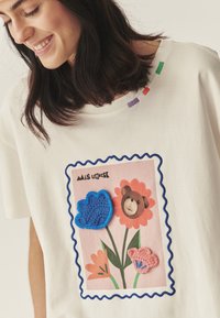 Biała bawełniana koszulka z kolorowym nadrukiem przedstawiającym niedźwiedzia i kwiaty, ozdobiona niebieskim falistym haftem oraz wielobarwnym przeszyciem.